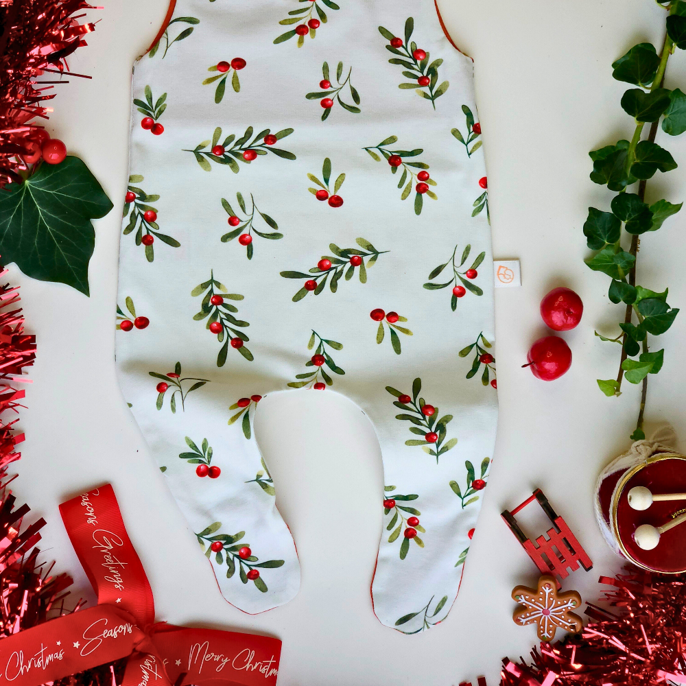 A baby jumpsuit laid flat on a white surface, decorated with a green mistletoe and red berry pattern. The jumpsuit has orange trim along the neckline and leg openings, and orange buttons at the shoulder straps.