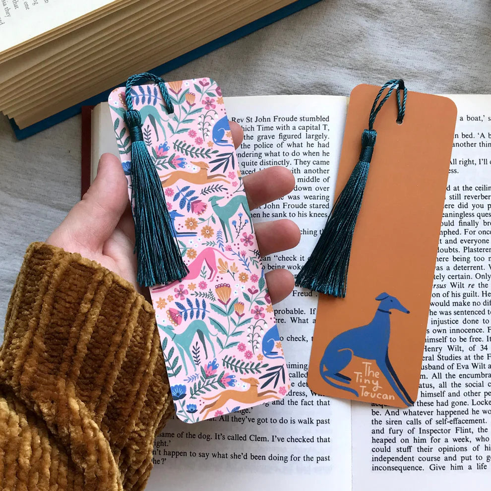 Hand holding two bookmarks, one floral and one with a dog design, over an open book.