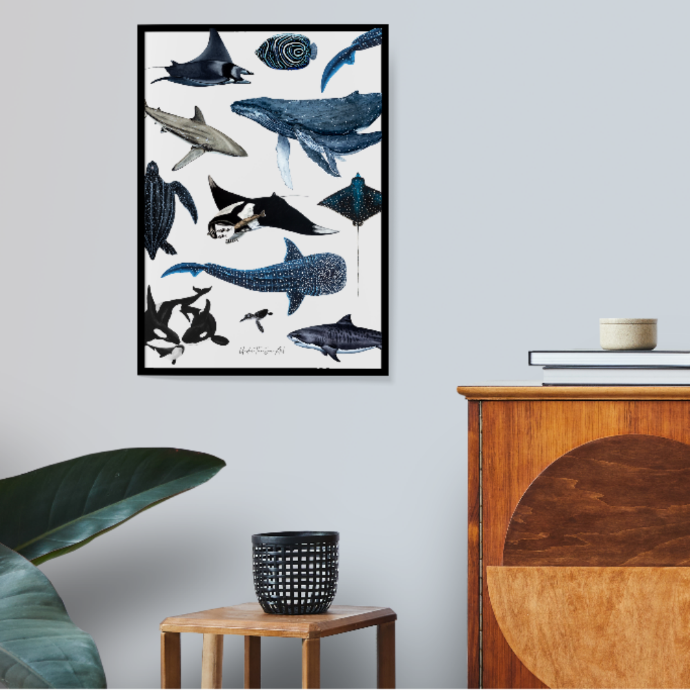 A framed vertical art print featuring various blue and gray whale and shark illustrations on a white background, hanging on a light gray wall in a styled room with a wooden cabinet, a small plant, and books.