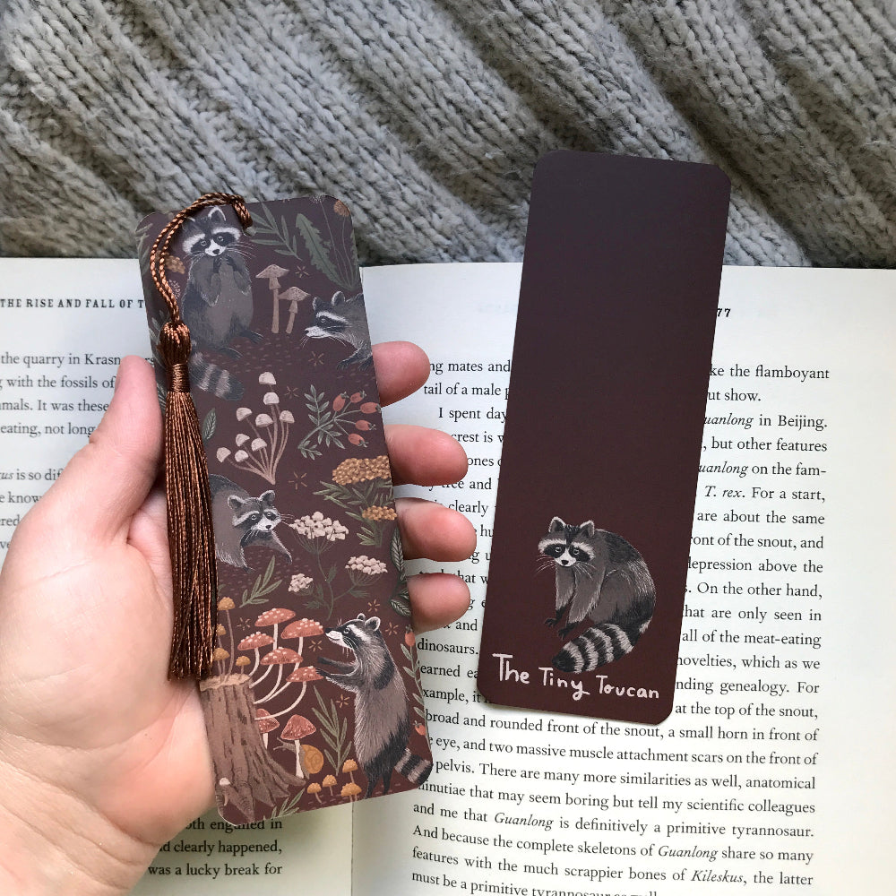 Hand holding a decorative bookmark with raccoon design on an open book.