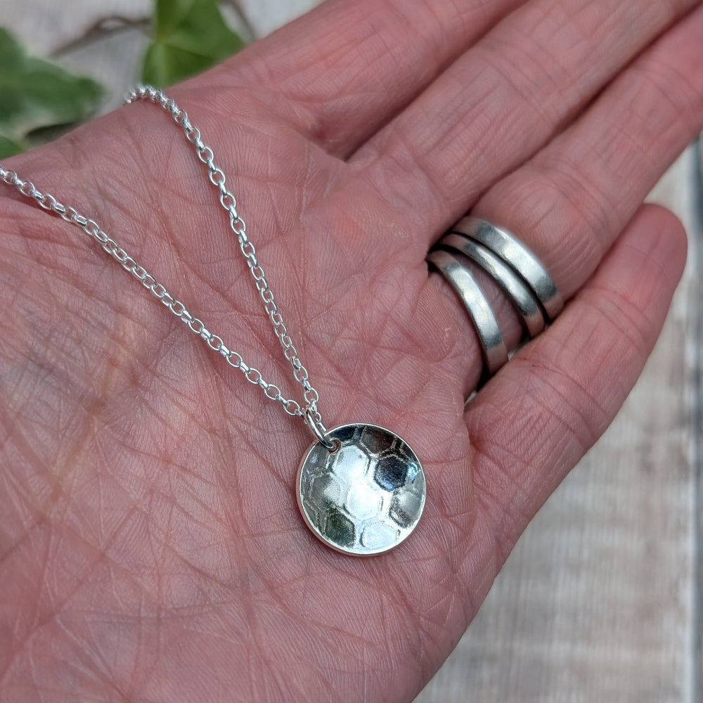 A silver pendant necklace with a hammered circular disc charm, displayed on a person's palm. The person is also wearing three silver bands on their fingers.