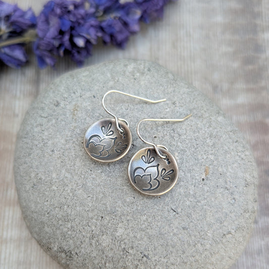 Silver earrings with floral designs on a stone surface with purple flowers in the background