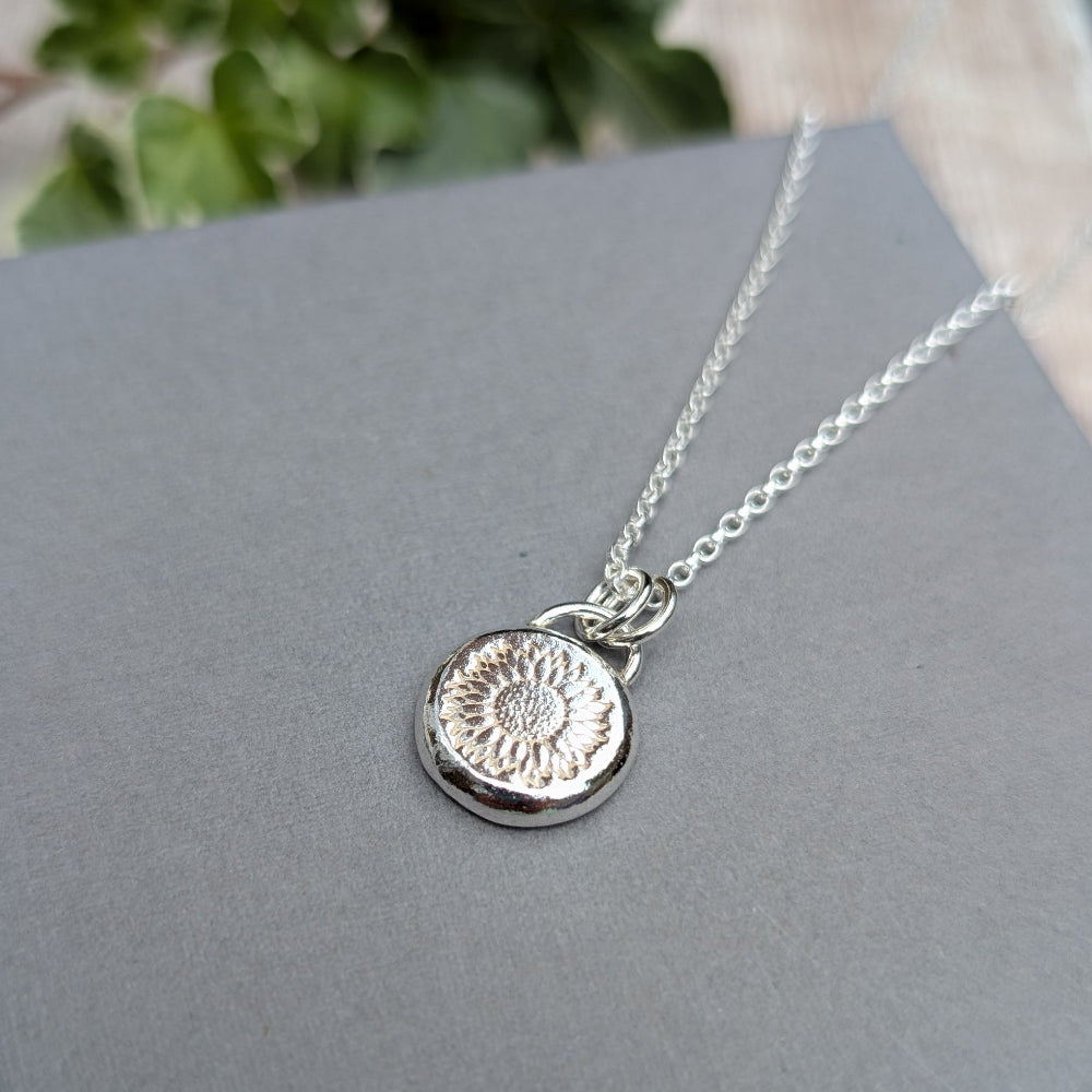 A sterling silver necklace with a textured circular pendant featuring a floral or sunburst pattern, resting on a gray surface.