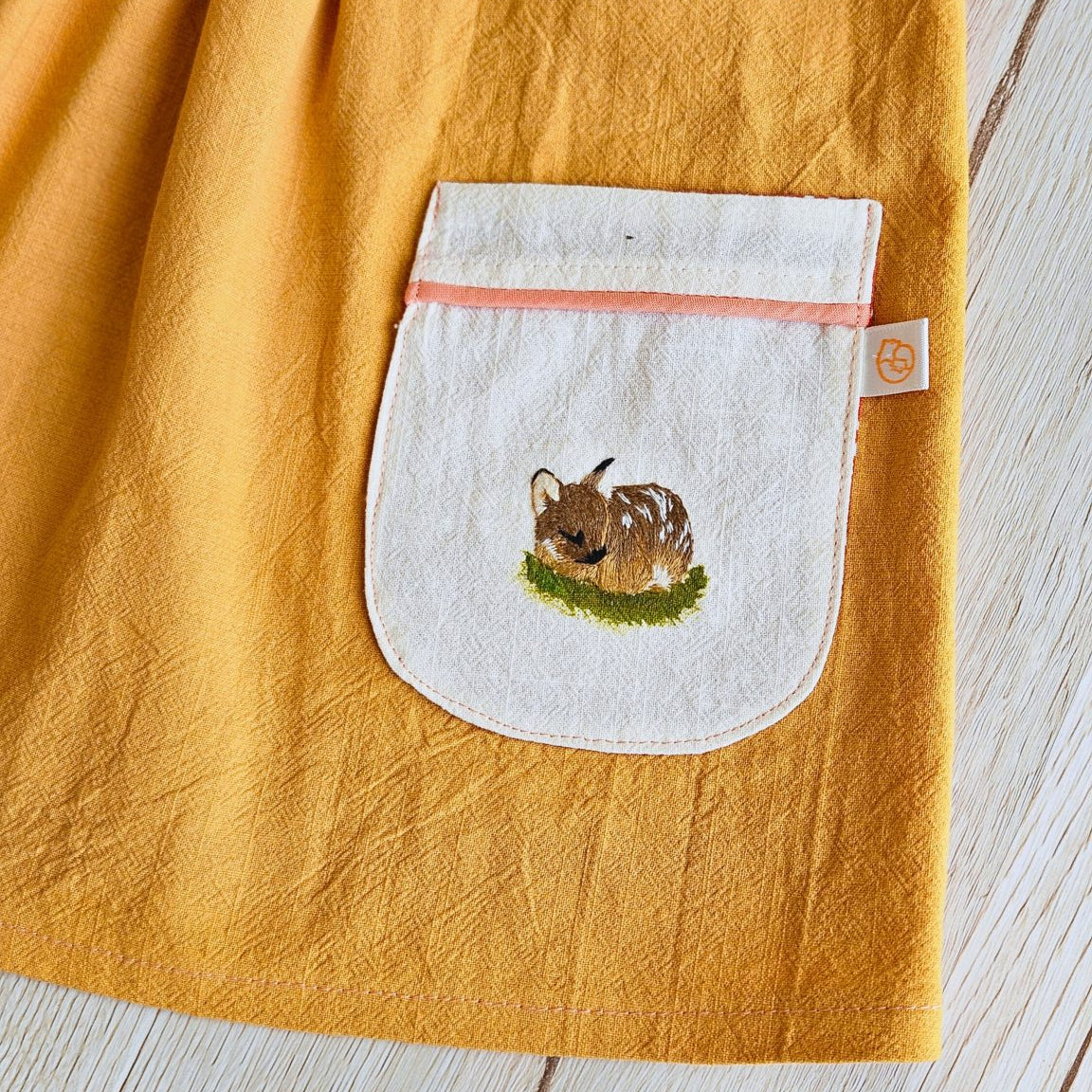 A close-up of a mustard yellow linen fabric item, likely a towel or blanket, featuring a white patch pocket with a small embroidered brown animal and green grass, laid flat on a light wood surface.