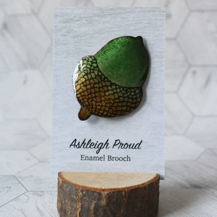 Green acorn enamel brooch on a card with 'Ashleigh Proud' branding, displayed on a wooden stand against a geometric patterned background.