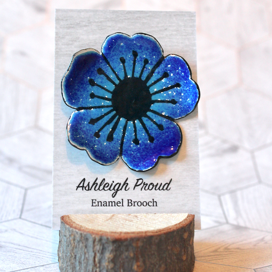 Blue flower-shaped enamel brooch on a card with 'Ashleigh Proud' branding, placed on a wooden block.