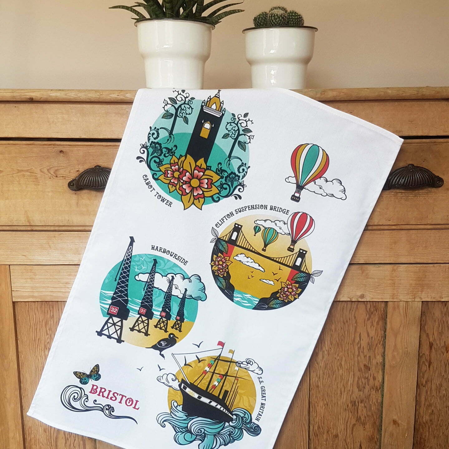 A white kitchen tea towel featuring colorful vintage-style illustrations of Bristol landmarks and symbols, draped over a wooden chest with potted plants in the background.