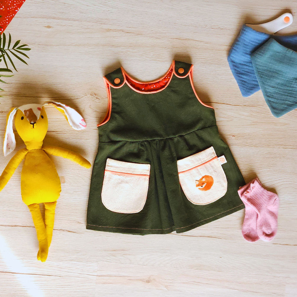 A styled flat-lay of a baby outfit set on a light wood floor, featuring an olive green pinafore dress with cream pockets, a blue and teal bib, pink booties, and a yellow plush bunny toy.