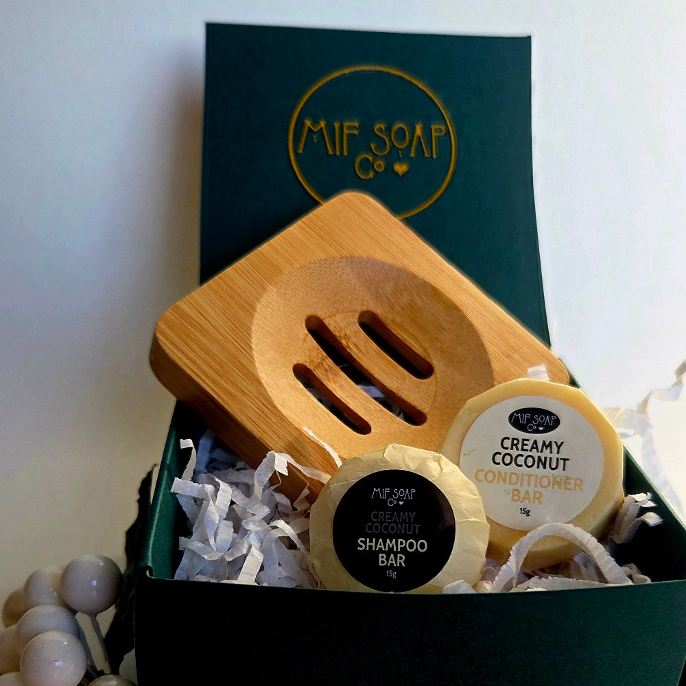 A Mif Soap Co gift set in a dark green box, featuring a bamboo soap dish, a shampoo bar & a conditioner bar, arranged with white paper filler and decorative pearls.