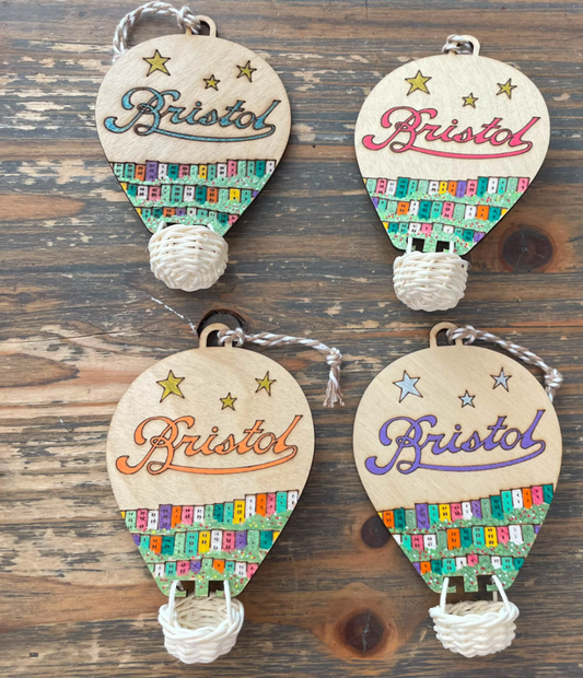 Four wooden hot air balloon ornaments arranged on a rustic wooden surface. Each ornament is made of natural wood with colorful patterned paper, silver string, and a white woven basket.