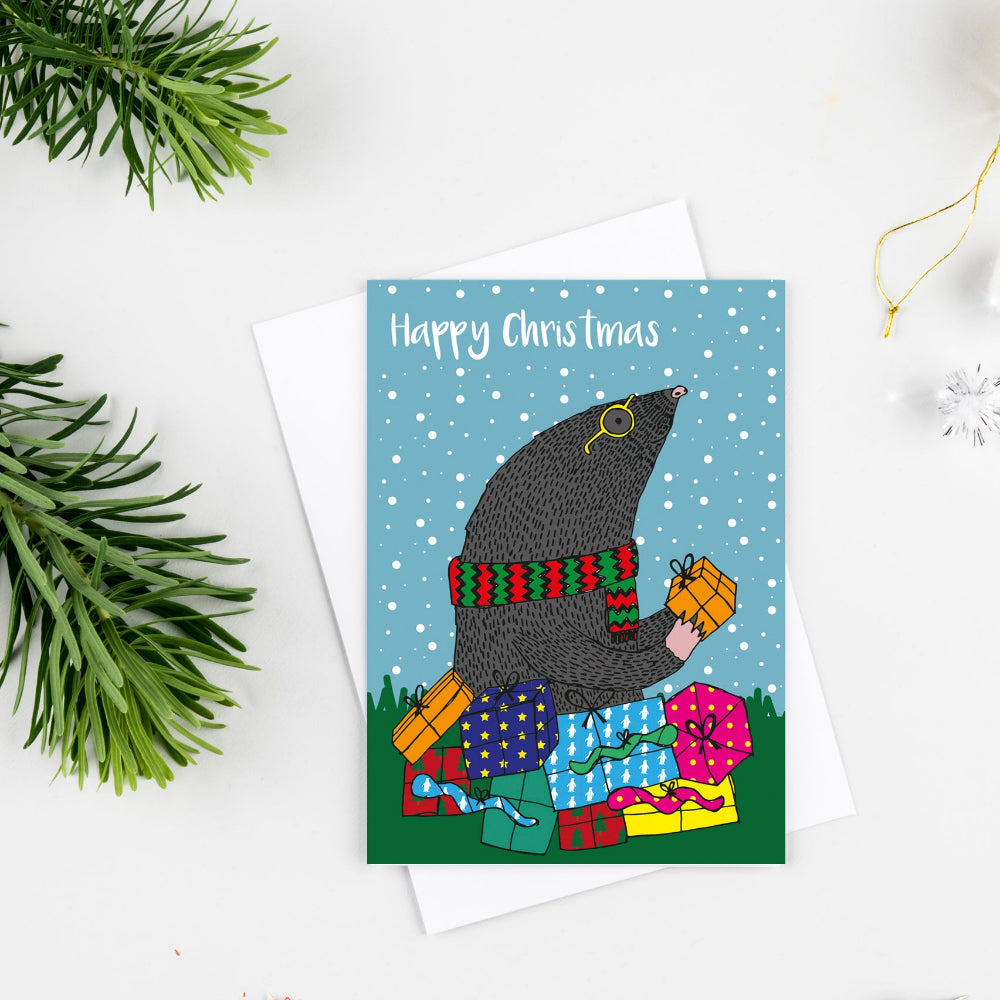 A Christmas greeting card featuring a moose wearing a witch hat and scarf while stacking presents, with the text 'Happy Chris Christmas' on a snowy blue background. The card is styled in a flat lay with a white envelope, green pine branches, a red pencil, and holiday ornaments on a white surface.