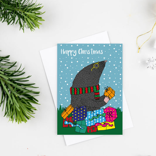 A Christmas greeting card featuring a moose wearing a witch hat and scarf while stacking presents, with the text 'Happy Chris Christmas' on a snowy blue background. The card is styled in a flat lay with a white envelope, green pine branches, a red pencil, and holiday ornaments on a white surface.