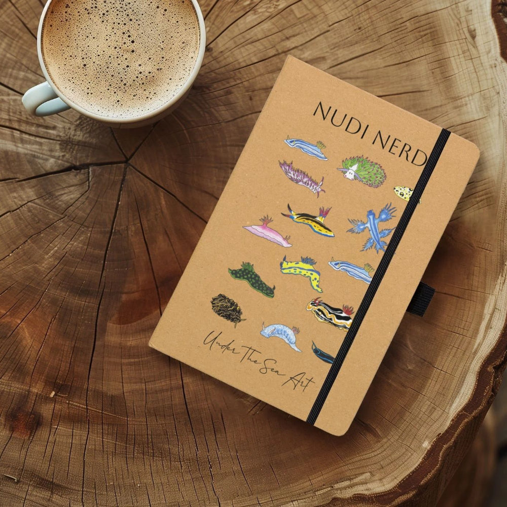 A tan hardcover notebook titled 'NUDI NERD' with colorful nudibranch illustrations and a black elastic closure, placed on a rustic wood slice table next to a cup of coffee.