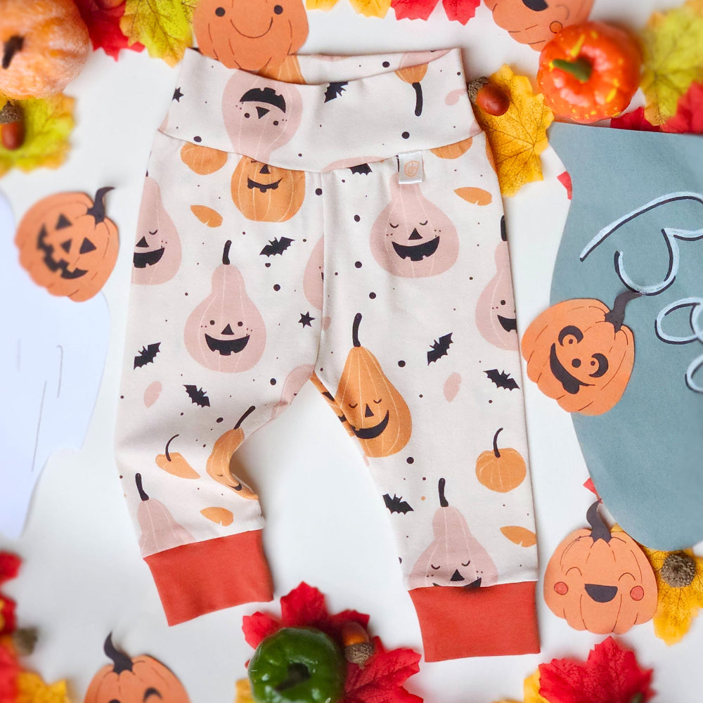 A pair of baby leggings with a Halloween print featuring pumpkins, gourds, bats, and ghosts on an off-white background, styled in a flat lay with autumn leaves, small pumpkins, and a blue blanket.