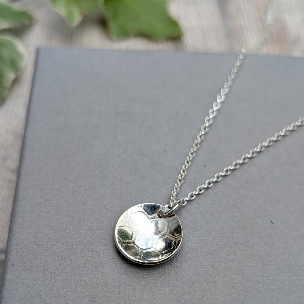 A silver disc pendant necklace with a textured, hammered finish on a delicate silver chain, displayed on a gray book with blurred green leaves in the background.
