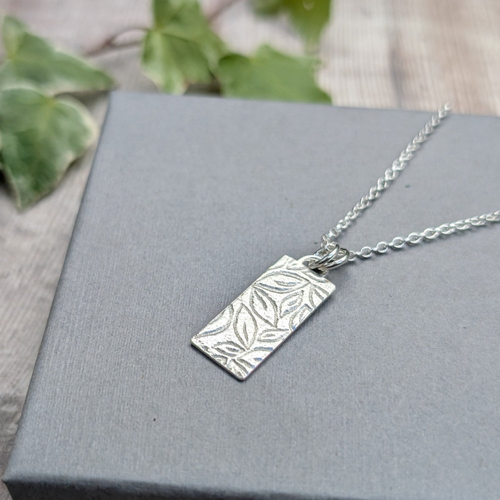 A silver rectangular pendant necklace with an embossed leaf pattern, resting on a gray textured box with blurred green leaves in the background.