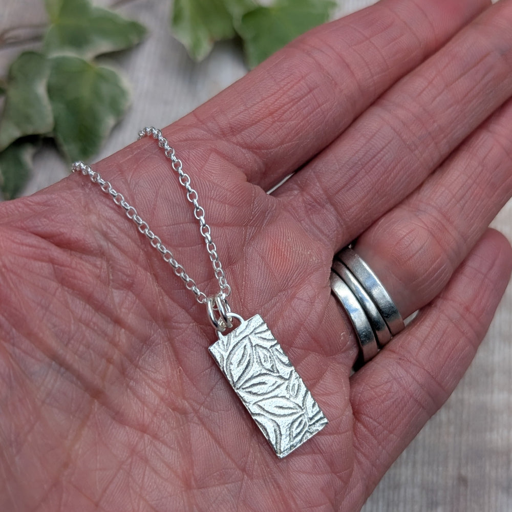 A silver rectangular pendant necklace with an embossed leaf pattern, resting on an open hand. The hand also has several silver rings on the fingers, and blurred green leaves are visible in the background.