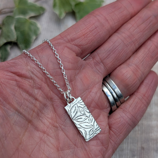 A silver rectangular pendant necklace with an embossed leaf pattern, resting on an open hand. The hand also has several silver rings on the fingers, and blurred green leaves are visible in the background.