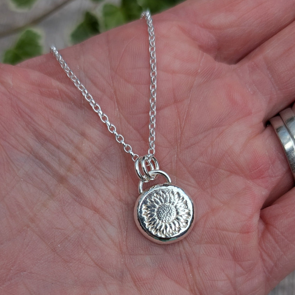 A sterling silver necklace with a round sunflower pendant is displayed on an open hand. The silver chain and pendant are clearly visible, with the textured sunflower design on the pendant highlighted by natural light.