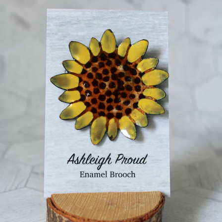 Sunflower-shaped enamel brooch on a card with 'Ashleigh Proud' branding, displayed on a wooden stand.