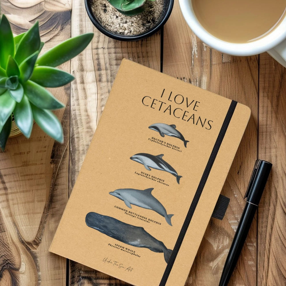 A tan hardcover notebook with black text reading 'I LOVE CETACEANS' and illustrations of dolphins and a whale, styled on a wooden tabletop with a plant, coffee, and a pen.
