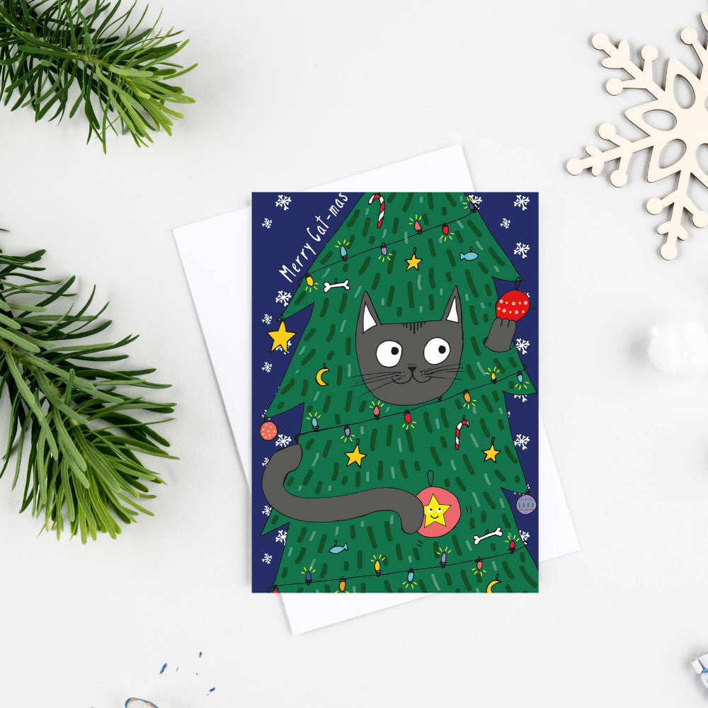 A Christmas greeting card featuring a black cat peeking out from behind a decorated green Christmas tree, styled in a flat lay with pine branches, a wooden snowflake, and stationery on a white background.