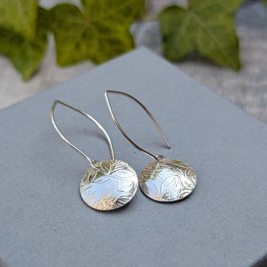 A pair of silver drop earrings with textured, hammered circular discs and curved wire hooks, displayed on a gray square block with blurred green leaves in the background.
