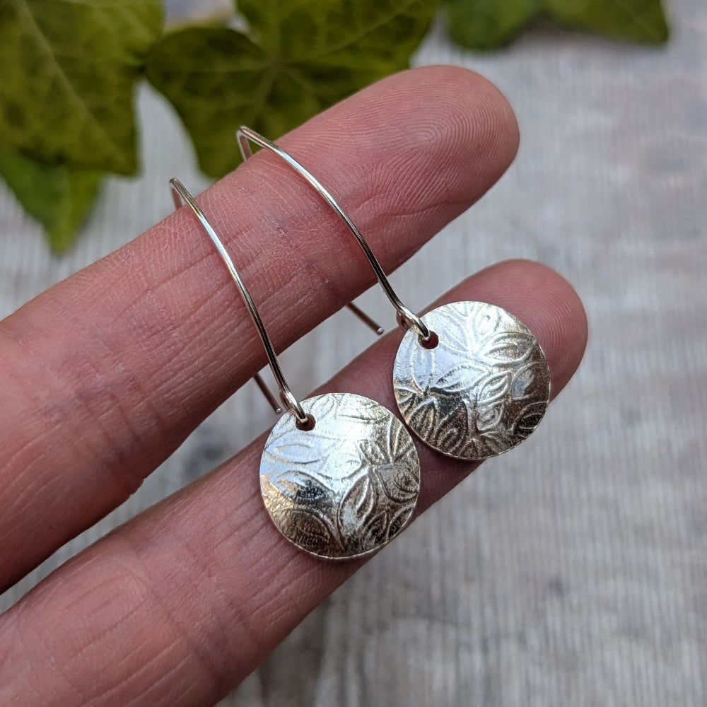 A pair of silver drop earrings with circular, textured disc charms held on a person's fingers against a light gray background with green leaves at the top.