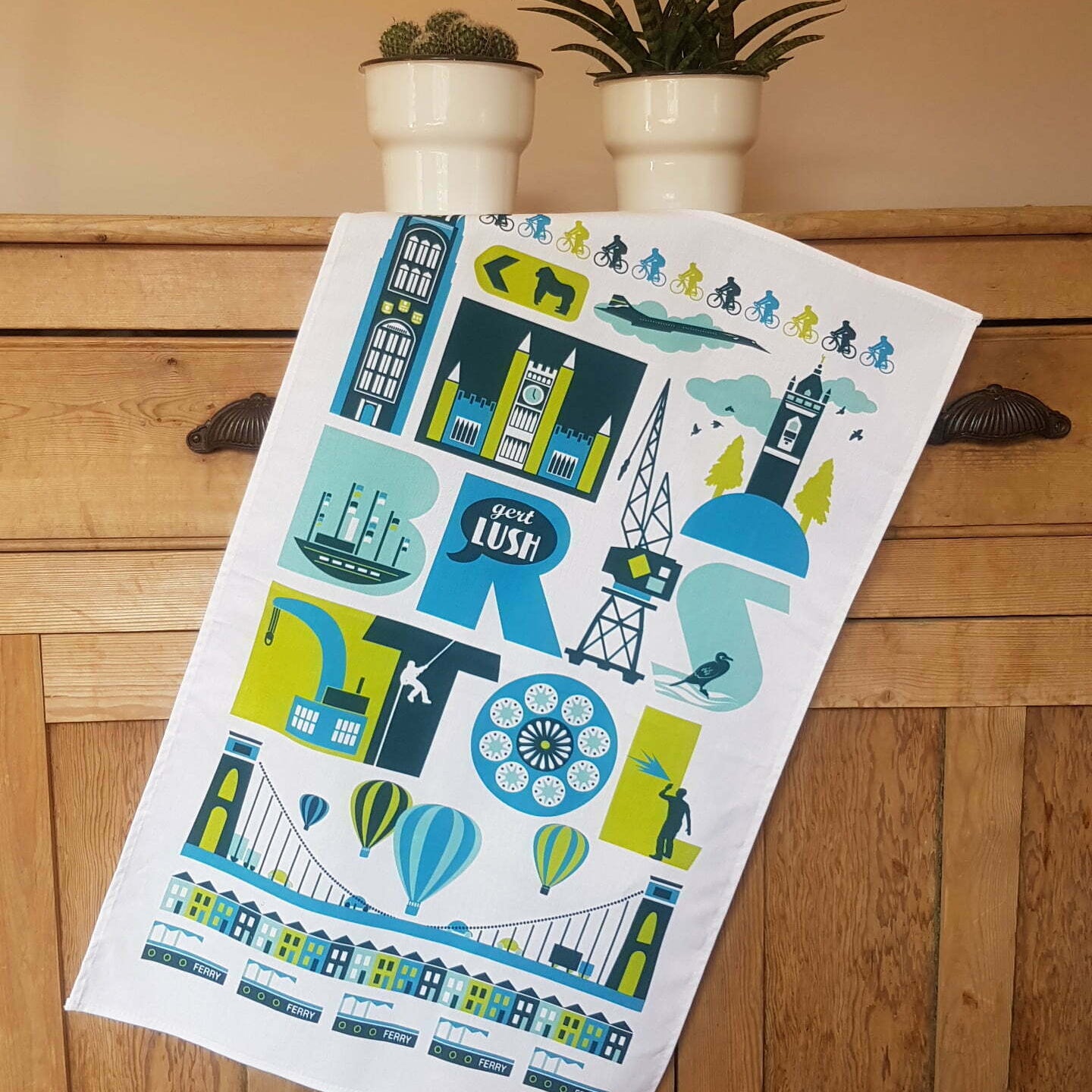 A white kitchen tea towel featuring a colorful graphic illustration of London landmarks and symbols, draped over a wooden chest with potted plants in the background.