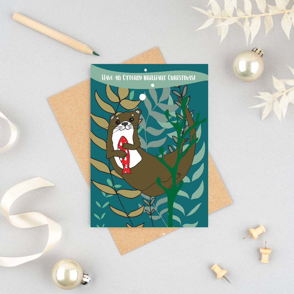 A Christmas greeting card featuring an illustration of an otter holding a red and white striped ornament, styled on a light gray surface with a kraft envelope, white ribbon, push pins, and pearl ornaments.